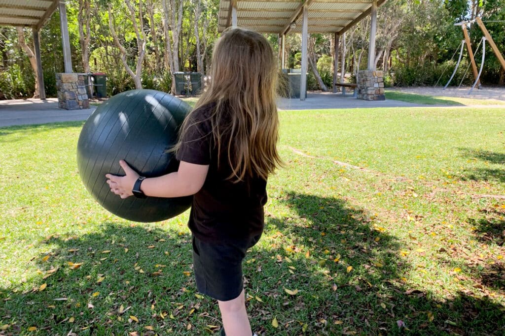 Child with long hair seen from behind carrying a large black exercise ball across a grassy park toward a shaded pavilion and playground. The quiet outdoor moment reflects how to start homeschooling by giving neurodivergent kids space to decompress, move at their own pace, and feel safe before structured learning begins.