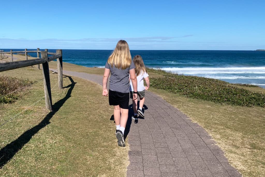 Two children walk side by side along a paved coastal path with ocean waves rolling in beside them and open sky ahead. The scene reflects starting homeschooling as a shared journey that can feel heavy at first while carrying change, but also allows space to move forward together at a gentler pace.